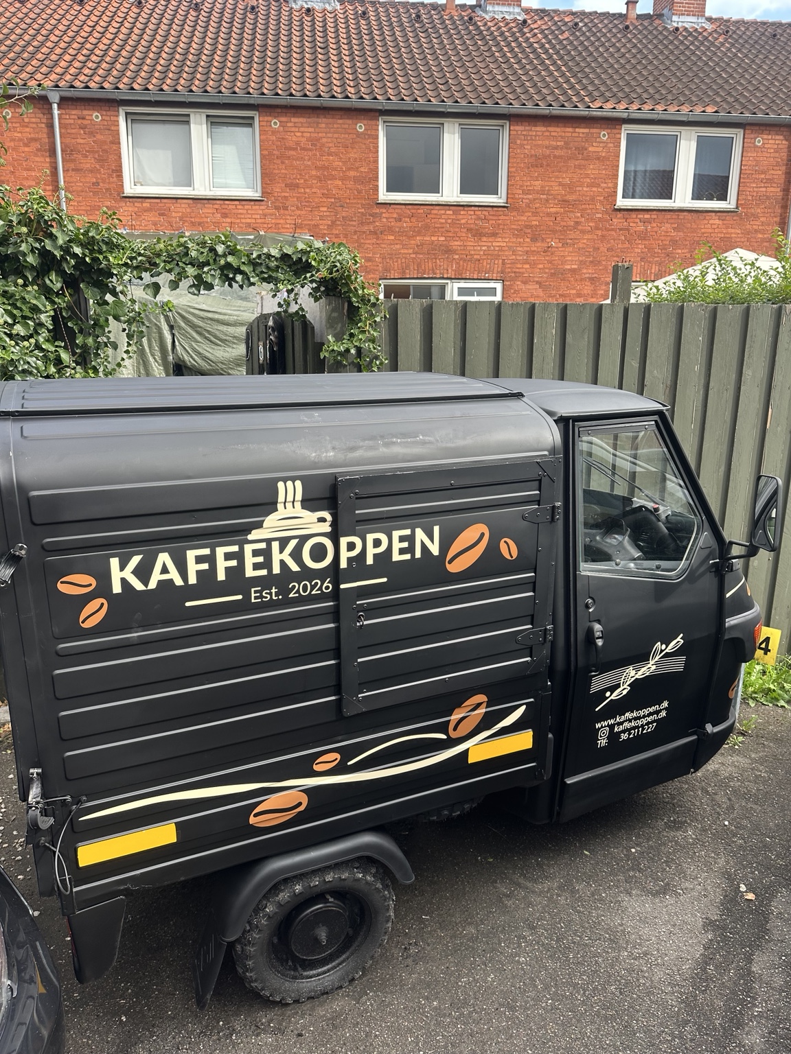 Kaffekoppens sorte tuk tuk med cremefarvet logo og orange kaffebønner på siden.
